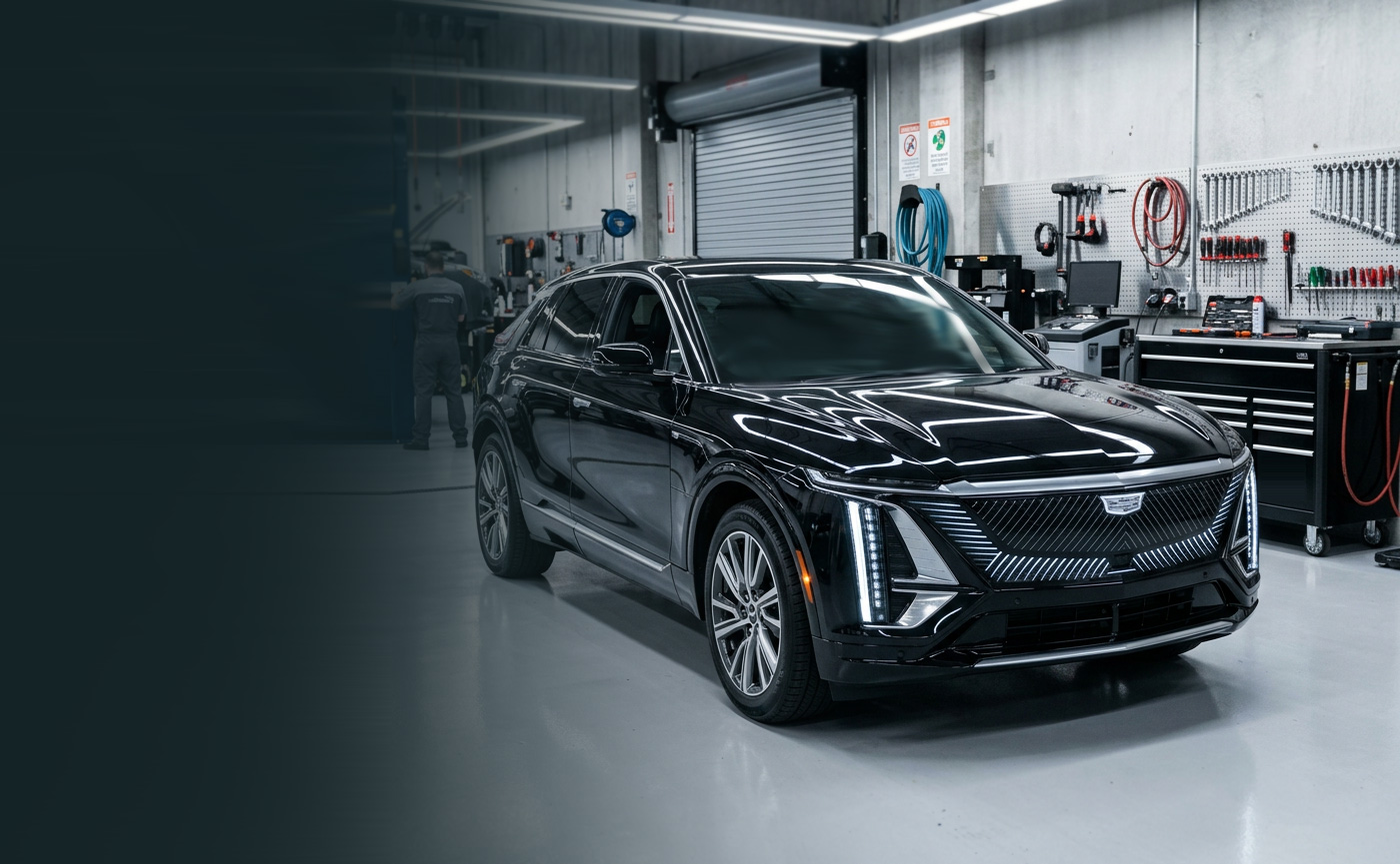 A black Cadillac Lyriq standing on a body-shop floor, a technician at the bench behind it