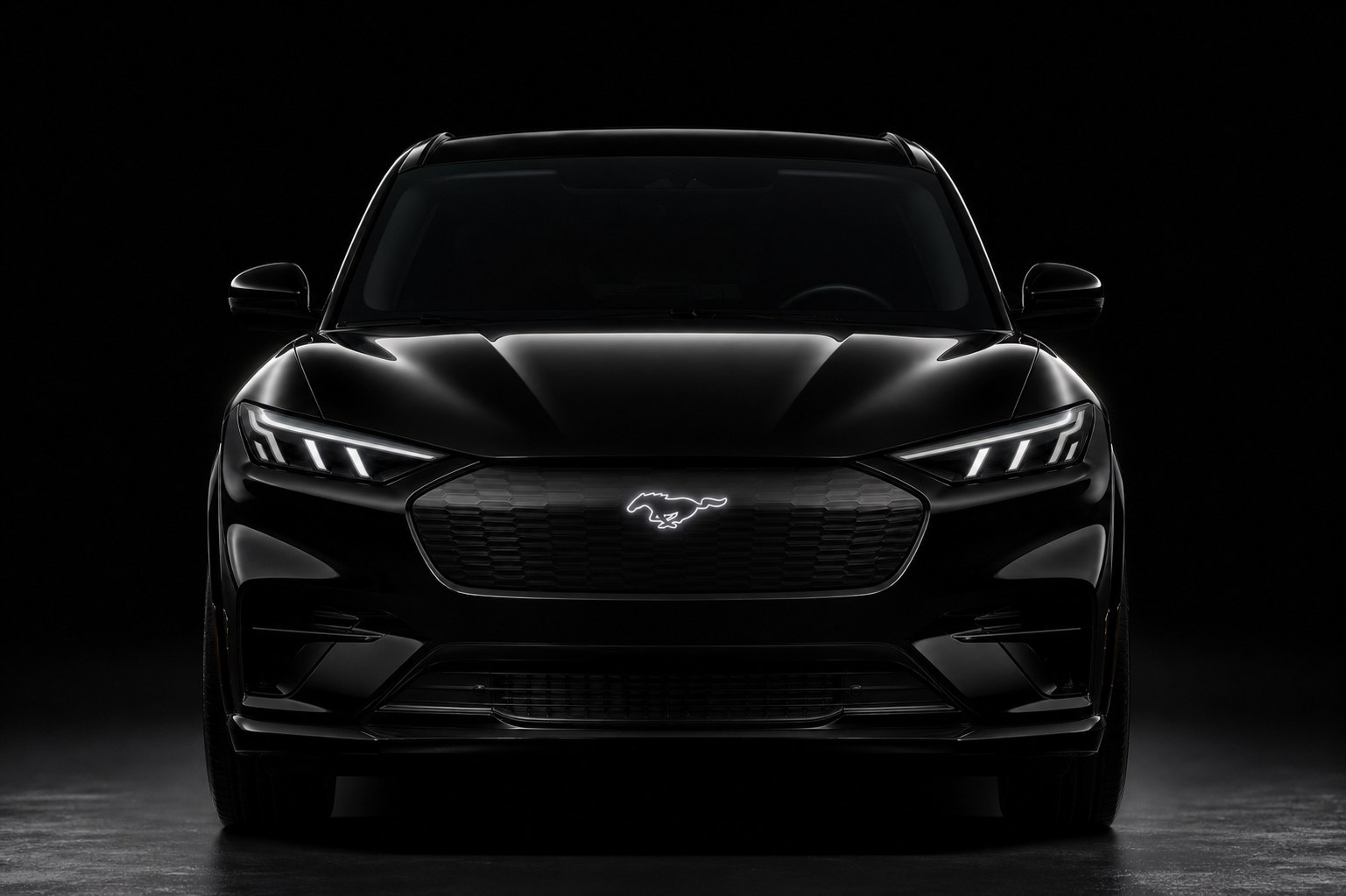 The front of a black Ford Mustang Mach-E in a dark studio: hood, LED headlights, the closed grille panel with the illuminated pony badge and the lower bumper — the panels a collision repair replaces
