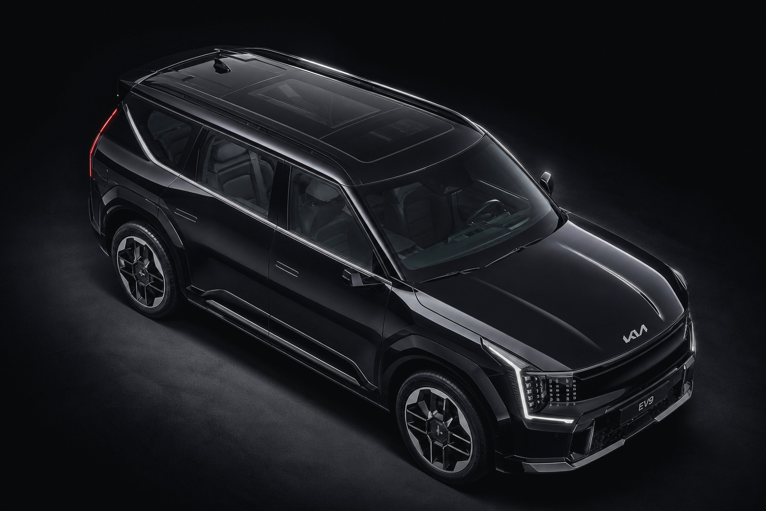 A black Kia EV9 seen three-quarter from above the front wing against a dark studio ground: the roof and its panoramic glass, the full flank, the hood, the headlight signature and the front bumper