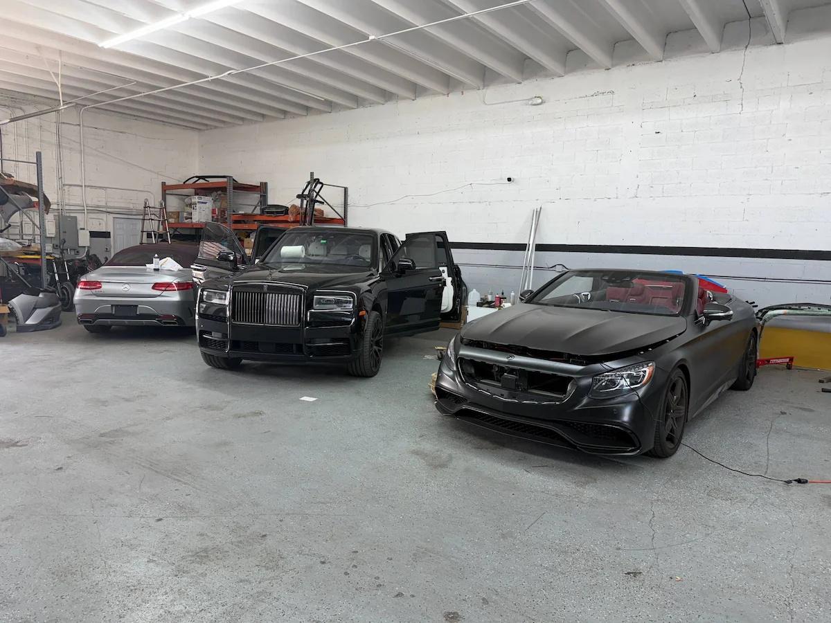 A black Rolls-Royce Cullinan beside a matte grey Mercedes-AMG cabriolet with its front bumper removed, standing inside the shop.