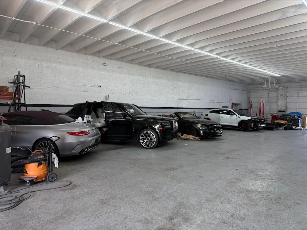 A row of cars along the shop wall with doors open and covers laid over the panels, waiting on refinishing work.