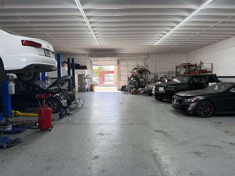 The length of the shop seen from the back, a car raised on the lift at the left and finished cars parked along the right-hand wall.