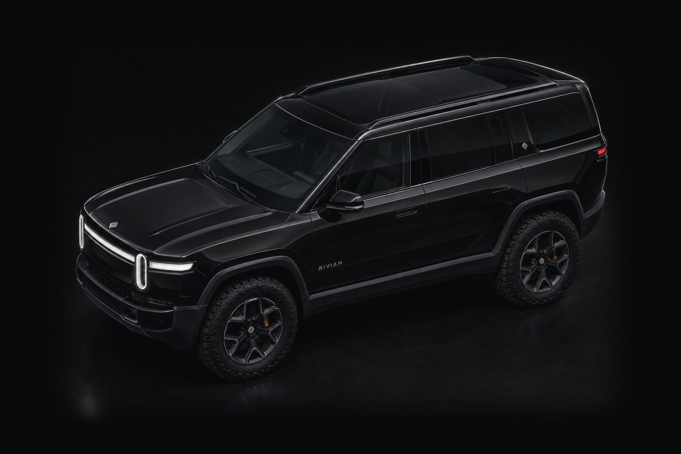 A black Rivian R1S seen three-quarter from above and ahead of the front wing against a dark studio ground